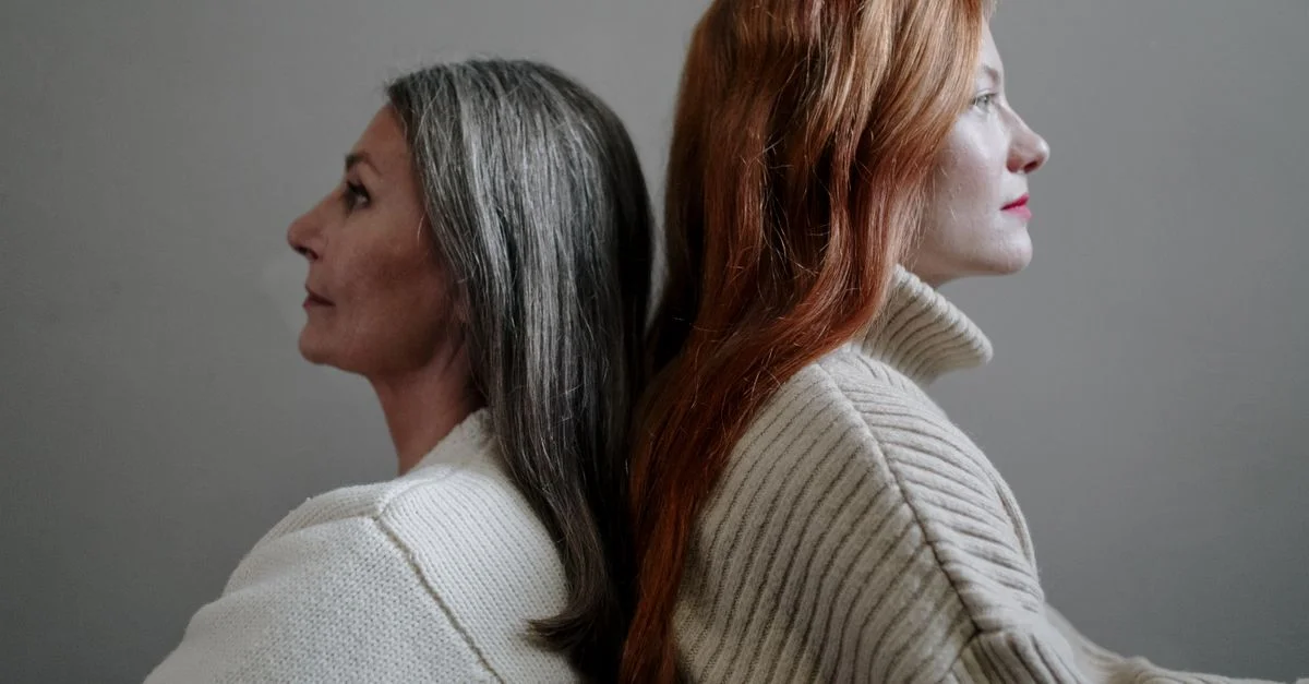 Une vue latérale en gros plan de deux femmes assises dos à dos dans des pulls tricotés, avec un fond neutre.