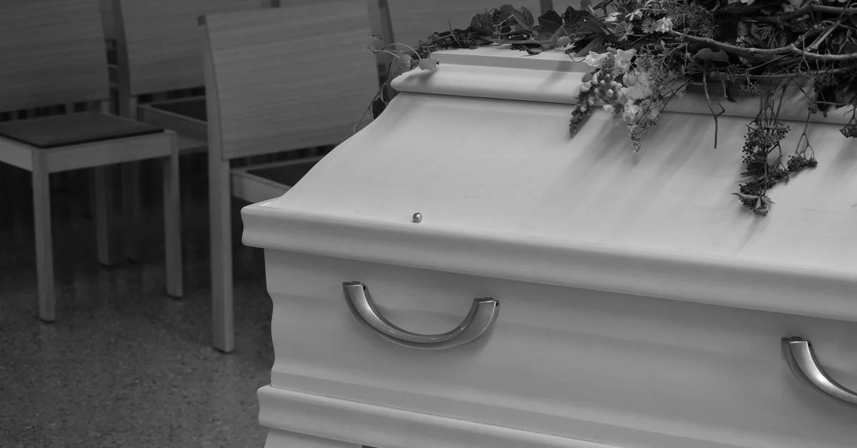 Image en niveaux de gris montrant un cercueil avec des fleurs dans une mise en scène funéraire, capturant une atmosphère de chagrin.
